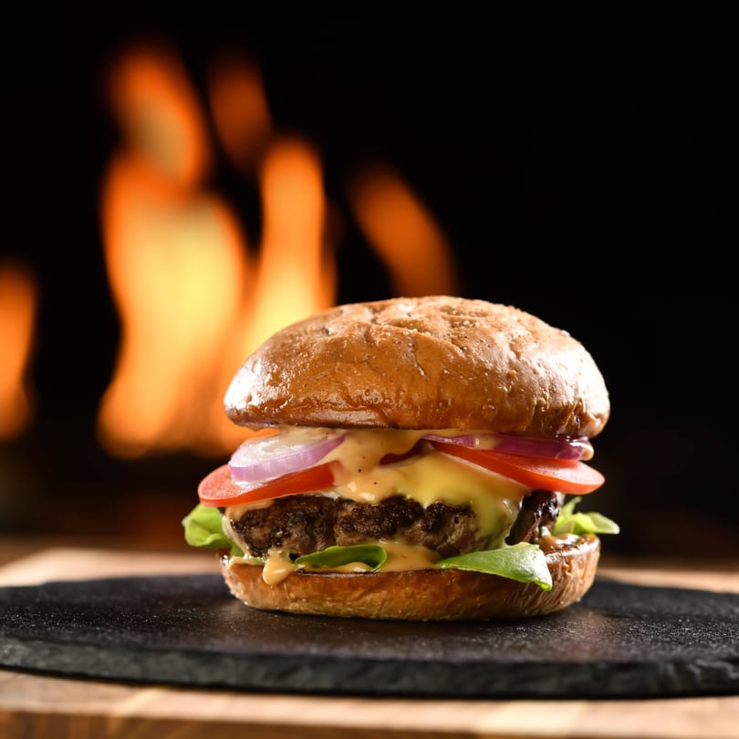 Gourmet burger with cheese, lettuce, tomato, and onion on a bun, with flames in the background.